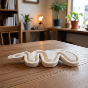 Könnte beinhalten: Ein cremefarbenes, schlangenartig geformtes Dekorationsobjekt liegt auf einem Holztisch. Das Objekt hat eine glatte, matte Oberfläche und ein erhabenes, wellenförmiges Design. Der Hintergrund umfasst ein Bücherregal, Pflanzen und eine Lampe.