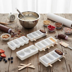 Könnte beinhalten: Weiße Eis am Stiel-Formen in verschiedenen Formen, darunter Rechtecke und Herzen, auf einer Holzoberfläche. Zutaten wie Nüsse, Beeren und Streusel befinden sich in kleinen Glasschalen. Eine Schüssel mit Teig, ein Schneebesen und eine Schokoladenflasche sind ebenfalls vorhanden.