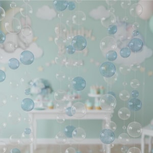Puede incluir: Guirnaldas de burbujas transparentes y azul claro cuelgan frente a una pared verde claro decorada con nubes y estrellas. Las burbujas varían en tamaño y están ensartadas en líneas transparentes, creando una atmósfera caprichosa y festiva. Una mesa blanca con cupcakes está al fondo.