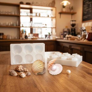 May include: A collection of seashells and white silicone molds in the shape of seashells. The molds are on a wooden surface, along with several real seashells of various sizes and colors, including a pink shell with a pearl.