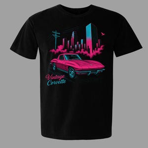 May include: Black t-shirt featuring a pink vintage Corvette graphic with a city skyline in the background. The design includes the text "Vintage Corvette" in a stylized font, with teal and pink accents. The car is a classic sports car.