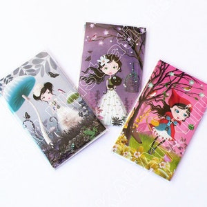May include: Three colorful notebooks with illustrations of girls in different settings. The first notebook features a girl under a blue mushroom, the second features a girl with a black cat, and the third features a girl in a red hooded cloak.