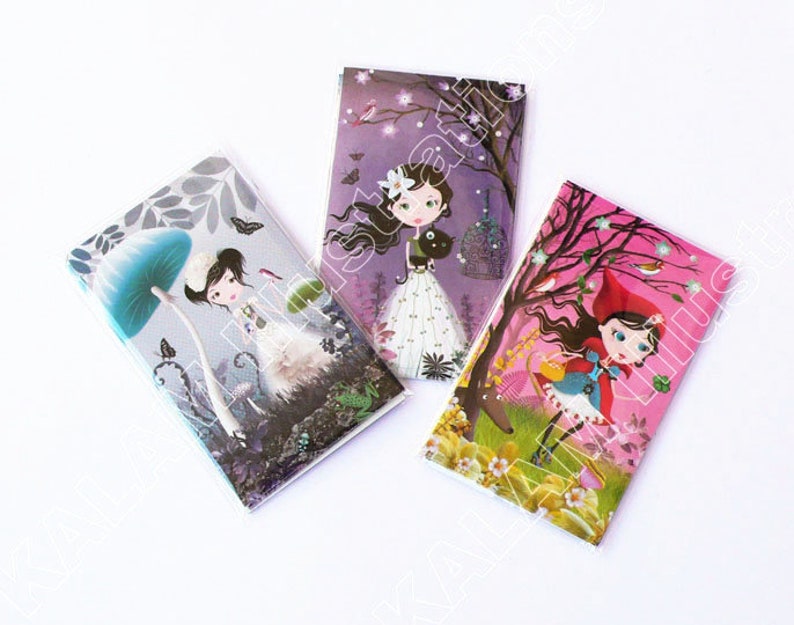 May include: Three colorful notebooks with illustrations of girls in different settings. The first notebook features a girl under a blue mushroom, the second features a girl with a black cat, and the third features a girl in a red hooded cloak.