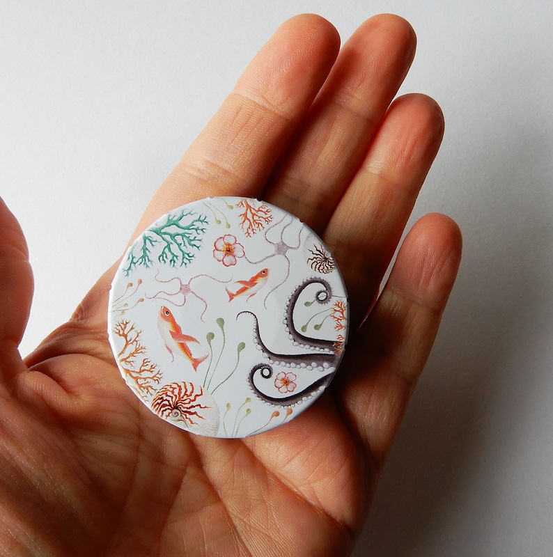 May include: A round, white compact with a colorful design featuring an octopus, coral, and fish. The design is reminiscent of an underwater scene.