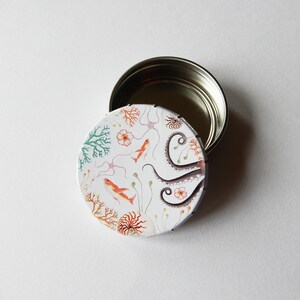 May include: A white round tin with a lid. The tin has a colorful design of an octopus, coral, and fish. The lid is silver.