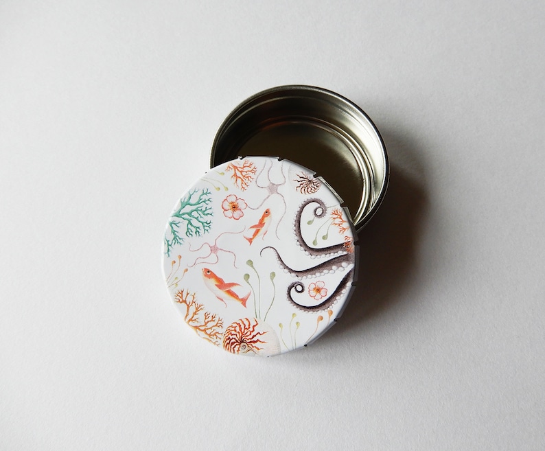 May include: A white round tin with a lid. The tin has a colorful design of an octopus, coral, and fish. The lid is silver.