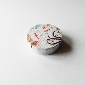 May include: A white round tin with a colorful design of an octopus, coral, and fish. The tin has a hinged lid and a scalloped edge.