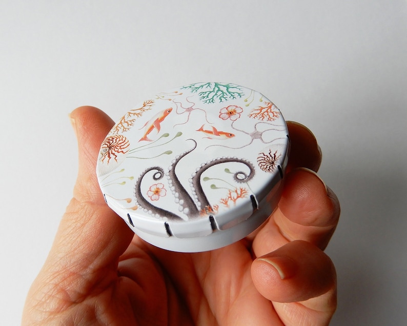 May include: A white round tin with a colorful design of an octopus, coral, and fish. The tin has a black stitched edge.