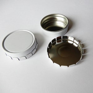 May include: A white metal tin with a scalloped edge. The lid is off and the inside of the tin is silver.