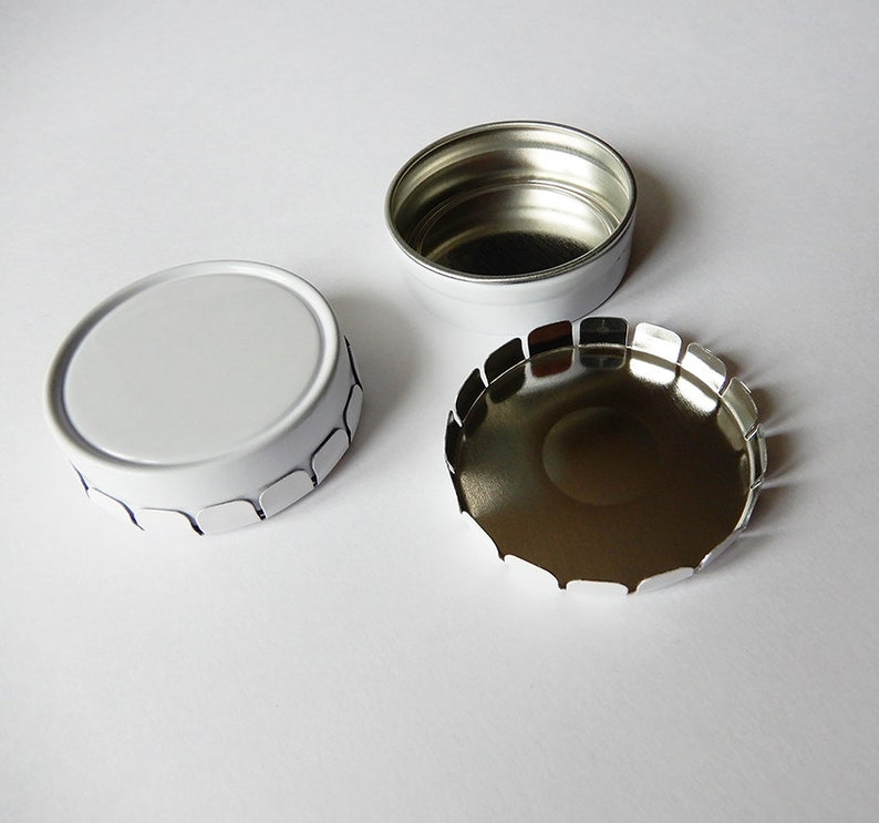 May include: A white metal tin with a scalloped edge. The lid is off and the inside of the tin is silver.