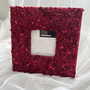 Peut inclure: Cadre photo carré rouge, orné de sequins et de perles. L'ouverture blanche est prévue pour une photo de 7,5 x 7,5 cm (3x3 pouces). Le texte "PHOTOFRAME..." est visible. Un objet décoratif pour afficher des photos.