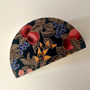 Puede incluir: Una caja decorativa semicircular con un fondo verde oscuro. La caja está adornada con un patrón de cerezas rojas, bayas azules y hojas amarillas y marrones. El diseño recuerda a una ilustración botánica.