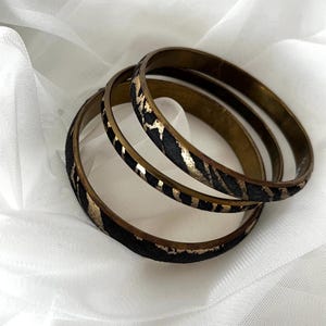 Könnte beinhalten: Drei Armreifen aus Messing mit einem schwarz-goldenen Tiermuster. Die Armbänder sind übereinander gestapelt und liegen auf einer weißen, strukturierten Oberfläche. Die Armreifen haben einen Vintage- oder Bohème-Stil.