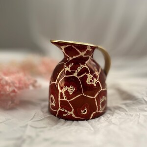 Può includere: Un piccolo brocca decorativa con una glassa rosso scuro e un bordo e un manico dorati. La superficie della brocca presenta un motivo astratto unico in oro, creando un contrasto sorprendente. Il design conferisce alla brocca un aspetto artigianale.