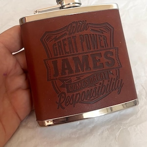 May include: A brown leather-wrapped stainless steel flask with a silver screw-top cap. The front features an embossed design with the text "With GREAT POWER JAMES COMES GREAT Responsibility" centred within a decorative frame and banner.