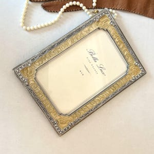 May include: A rectangular Bella Lux picture frame with a silver-toned metal border, cream-coloured enamel inlay featuring a swirling pattern, and small clear crystals. The insert reads Bella Lux Fine Frames 4x6. A pearl necklace and brown leather gloves.