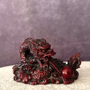 Peut inclure: Figurine de dragon rouge détaillée avec des écailles complexes et un corps recourbé. Le dragon est posé avec la tête levée, et une sphère rouge est à proximité. La figurine est sur une surface blanche.