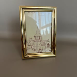 May include: A rectangular gold-toned metal picture frame containing a print of a church. The print features a brown line drawing of St. John the Baptist Church in Greatham, with the text St. John the Baptist Church Greatham printed at the top.