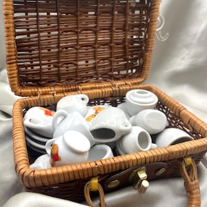 Puede incluir: Un juego de té en miniatura en una cesta de mimbre marrón. El juego incluye pequeñas tazas, platillos, una crema y un azucarero de porcelana blanca. Algunas piezas tienen diseños florales o de osos de peluche de colores. La cesta tiene un asa y un cierre.
