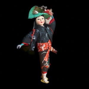 Peut inclure: Poupée japonaise vêtue d'une tenue traditionnelle, sur fond noir. La poupée porte un kimono noir avec des détails rouges, un obi rouge et un pantalon à motifs. Un chapeau vert en forme d'éventail orne la tête de la poupée. Le visage de la poupée est peint.