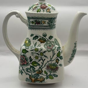 May include: A white ceramic teapot with a lid, handle, and spout. The teapot features a floral design in green, pink, yellow, and brown. The lid has a small knob, and the handle is curved.