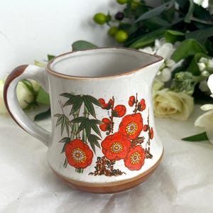 Op de afbeelding: Een keramieken melkkannetje met een crèmekleurige basis en een bruine rand en onderkant. De buitenkant is voorzien van een decoratieve print van groene bamboestengels en felrode bloemen met gele harten. Het heeft één handvat en een schenktuit.