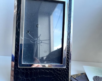 May include: A rectangular photo frame with a silver metal border and a black, textured, faux-leather insert. The frame is empty, with a glass pane. A small notebook and a card are visible in the background.