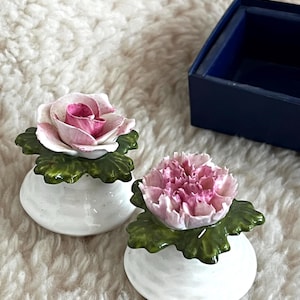 May include: Two white ceramic flower figurines with pink and white petals and green leaves. They sit on a white, fluffy surface. A dark blue box is in the background.
