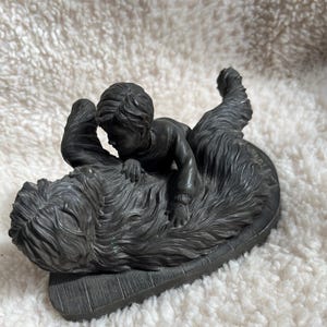 May include: A dark grey sculpture of a child riding a shaggy dog. The child is lying on the dog's back, with the dog's head and body forming the base of the sculpture. The sculpture is set on a rectangular base, and is made of a dark grey material.