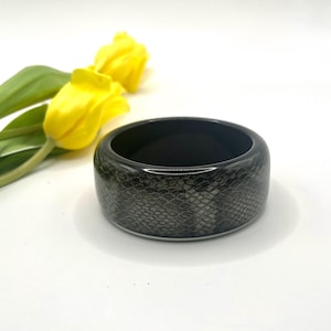 Könnte beinhalten: Ein breites, dunkelgraues Armreif-Armband mit Schlangenmuster. Das Armband befindet sich vor einem weißen Hintergrund, mit gelben Tulpen im Hintergrund.