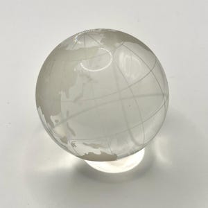 May include: A transparent glass globe featuring etched continents and grid lines. The globe rests on a clear, circular base, creating a subtle shadow. The world map is visible, displaying the Earth's landmasses.