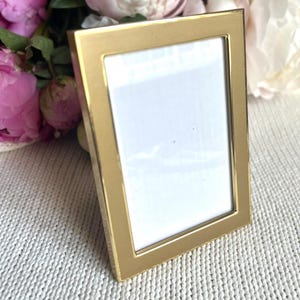 Puede incluir: Un marco de fotos rectangular dorado con interior blanco, ligeramente inclinado. Se encuentra sobre un fondo de tela blanca tejida y peonías rosas y blancas. El marco es de metal.