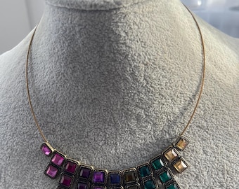 Op de afbeelding: Een ketting- en armbandset met vierkante, veelkleurige edelstenen in roze, paarse, blauwe, groene en gouden tinten. De sieraden hebben een bronzen metalen zetting en worden gepresenteerd op een grijze mannequin.
