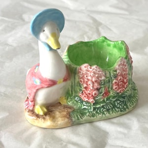 Könnte beinhalten: Ein Eierbecher aus Keramik mit einer weißen Ente, die eine blaue Haube und einen rosa Schal trägt. Die Ente steht neben einem grünen, strukturierten Sockel, der mit rosa und roten Blumenmustern verziert ist. Das Stück hat eine glänzende Glasur.