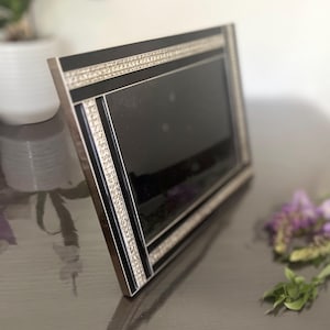 Op de afbeelding: Een rechthoekige fotolijst met een zwarte rand, geaccentueerd door twee parallelle stroken van heldere, sprankelende strass-steentjes. De lijst heeft een metallic zilverkleurige buitenrand en een donker, reflecterend glazen middenpaneel.