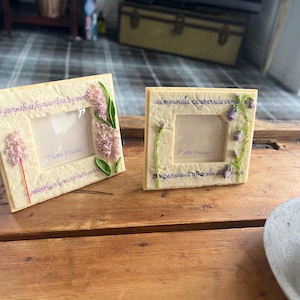 Può includere: Due cornici quadrate color crema con disegni floreali. Una cornice presenta un design di giacinto rosa, l'altra un design di campanula viola. Entrambe le cornici hanno le parole "Photo Frame" e testo botanico.