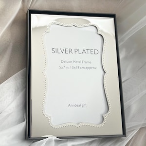 May include: Silver-plated deluxe metal frame, 5x7 inches, 13x18 cm approx. The frame has a decorative, scalloped inner edge and a black border. The text "SILVER PLATED" and "An ideal gift" are printed on the white background.