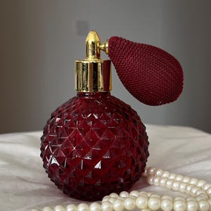 Può includere: Un flacone di profumo rotondo in vetro rosso scuro con un motivo a diamante testurizzato. Il flacone presenta un colletto in metallo color oro lucido e un ugello abbinato, con una pompetta atomizzatrice rivestita in tessuto rosso scuro.