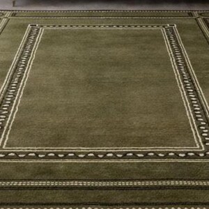 Olive green modern area rug with geometric border design placed in a minimalist living room with beige sofa and natural light