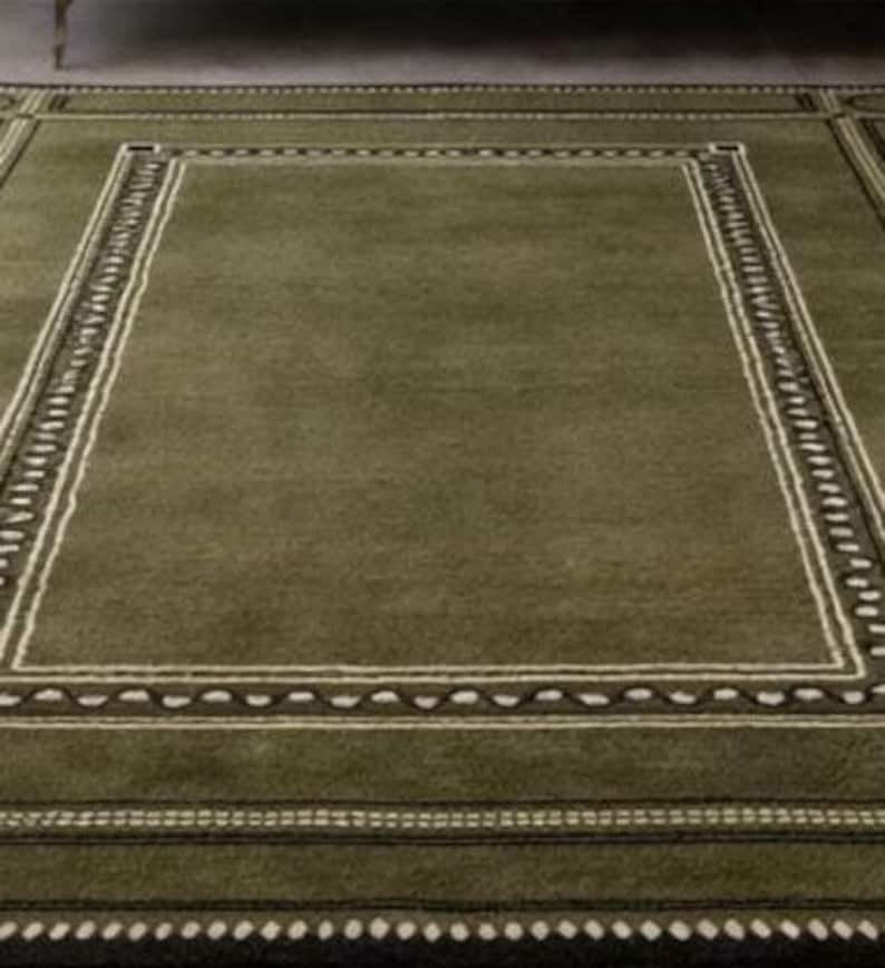 Olive green modern area rug with geometric border design placed in a minimalist living room with beige sofa and natural light