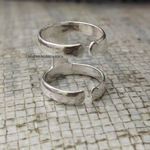 May include: A silver ring with an open design. The ring has two bands connected by a vertical bar. The text "SILVERRINGARTISANCO" is visible on the ring. The ring is displayed on a textured surface.