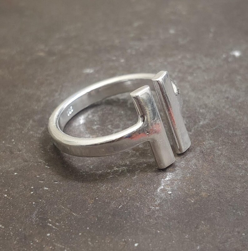 May include: A silver ring with a modern design. The ring features a band and two vertical rectangular bars that form a T-shape. The ring is made of a shiny, metallic material and is set against a dark, textured surface.