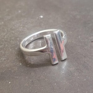 May include: A silver ring with a modern design. The ring features two vertical bars connected to the band, creating an open, geometric shape. The ring is made of a shiny, metallic material.