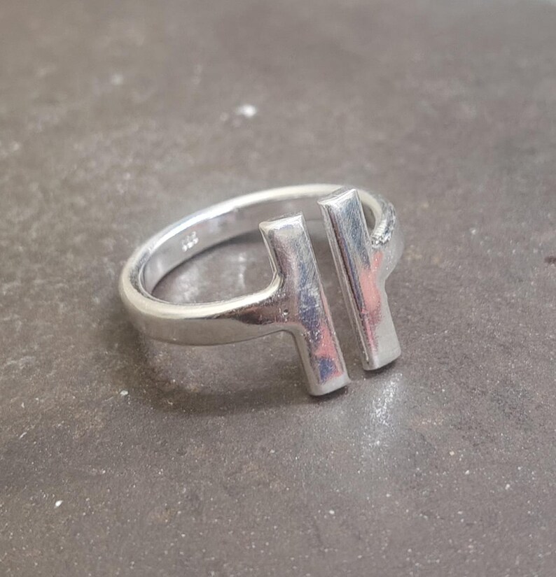 May include: A silver ring with a modern design. The ring features two vertical bars connected to the band, creating an open, geometric shape. The ring is made of a shiny, metallic material.