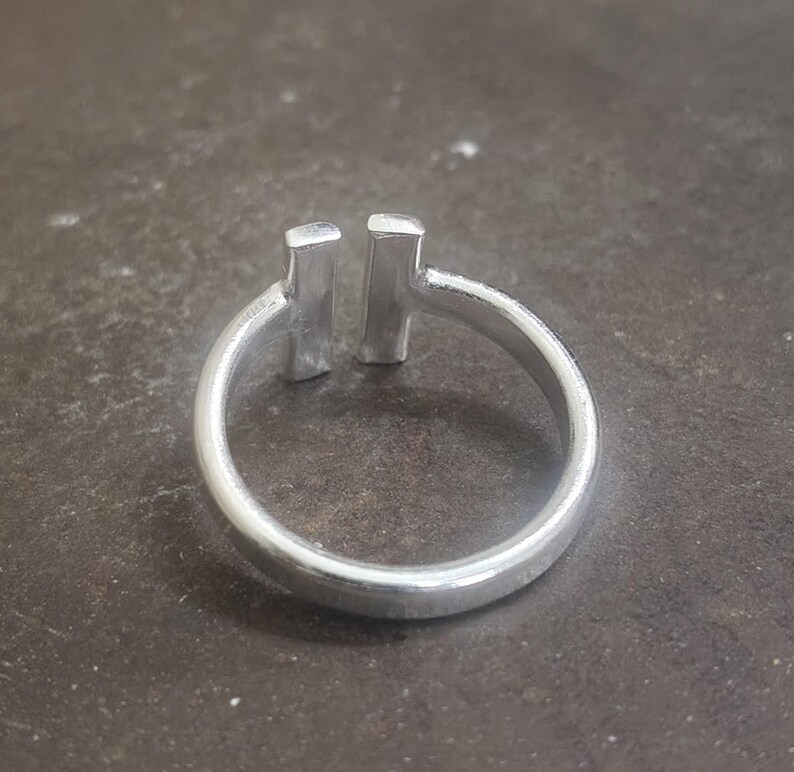 May include: A silver ring with a minimalist design. The ring features a smooth, rounded band and two vertical rectangular bars set close together at the top. The ring is made of a shiny, metallic material.