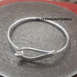 Peut inclure: Un bracelet jonc argenté avec une fermeture à crochet et œillet. Le bracelet est de forme circulaire avec une finition lisse et polie. Le texte "SILVERRINGARTISANCo" est visible.