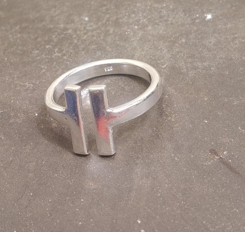 May include: A silver ring with a modern design. The ring features a band and a vertical bar design. The ring is made of a shiny, metallic material. The ring is shown on a gray surface.