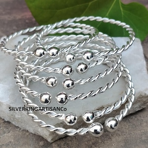 Può includere: Una collezione di braccialetti argentati in filo ritorto. Ogni braccialetto presenta un design a spirale e una piccola perla rotonda all'estremità. I braccialetti sono impilati su una superficie di pietra chiara. Il testo "SILVERRINGARTISANCo" è visibile.