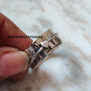 Peut inclure: Une bague spinner en argent avec un accent en forme de cœur. La bague a une bande texturée avec un motif floral. Le texte "SILVERRINGARTISANCo" est visible sur la bague. La bague est tenue par une personne.