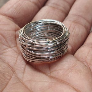 Puede incluir: Un anillo plateado con un diseño de alambre tejido. El anillo es grueso y tiene un aspecto texturizado, con múltiples capas de alambre que crean un aspecto artístico único. El anillo se muestra en una mano.